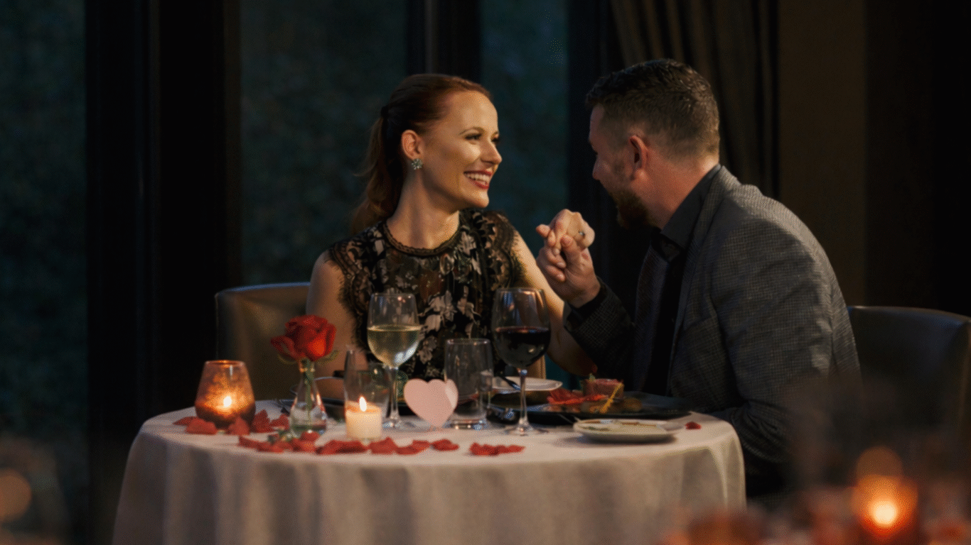 A couple sits at a round table set with wine, candles, a rose, and rose petals, smiling and holding hands in a dimly lit restaurant.