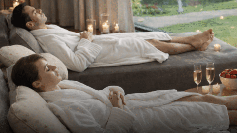 A man and a woman in white robes lie on lounge chairs indoors near a window, surrounded by candles, with champagne and strawberries on a nearby table.