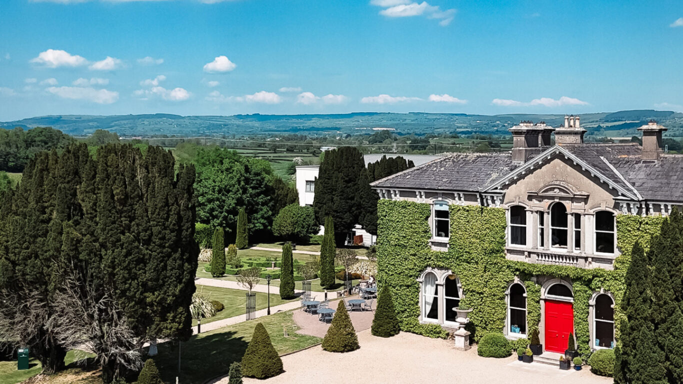 Elegant country hotel with ivy fa&ccedil;ade, red door, lush gardens, and scenic countryside views under a bright blue sky.