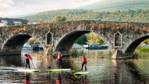 Guests paddleboarding on a scenic river under a historic bridge, surrounded by lush hills and moored boats, ideal for adventure.