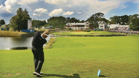 Guest plays golf on lush green course, with scenic views of the hotel, vibrant garden, and tranquil lake under a blue sky.
