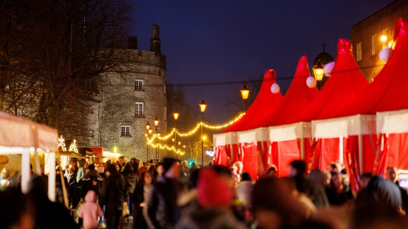 Guests enjoy a festive market with red tents, warm lights, and a historic stone tower, creating a lively evening atmosphere.